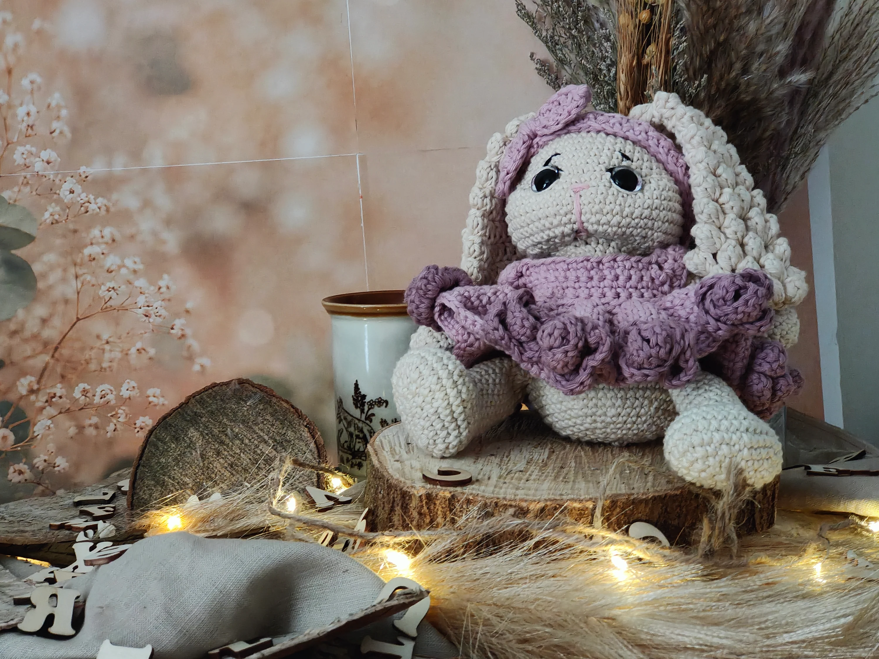 A handmade crochet bunny with purple clothing sitting on a wooden slice photo stage with warm fairy lights, dried flowers, and a cosy handmade backdrop.