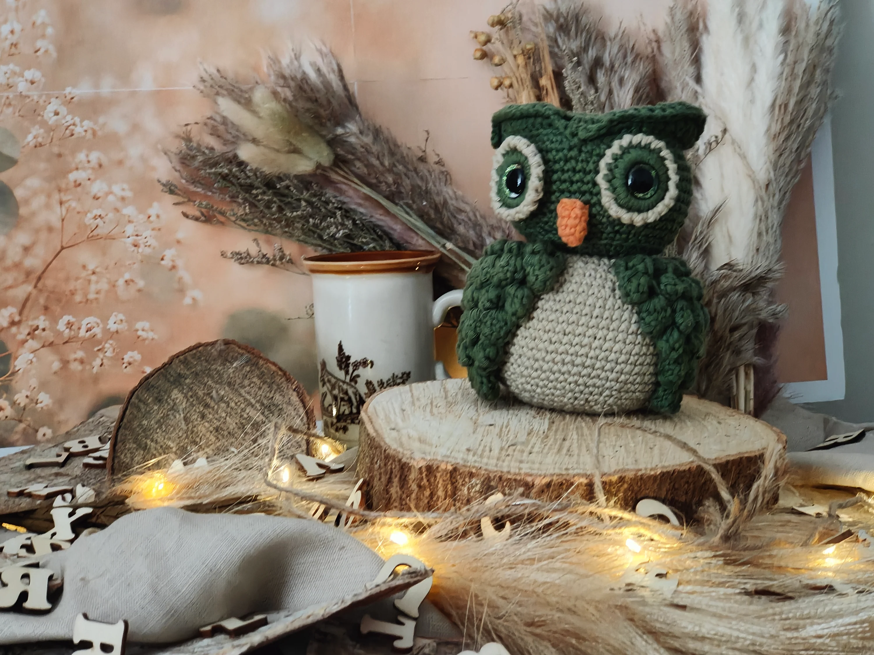 A handmade green crochet owl sitting on a wooden slice photo stage with warm fairy lights, dried flowers, soft fabric, and a cosy handmade backdrop.